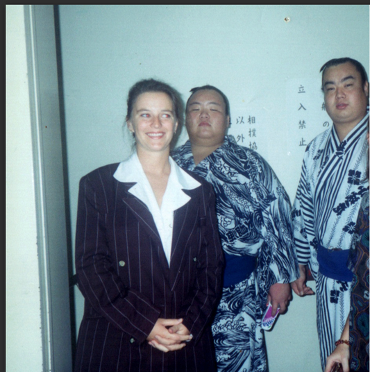 A woman in her twenties in Japan, smiling with a mix of happiness and nerves while standing between two sumo wrestlers.
